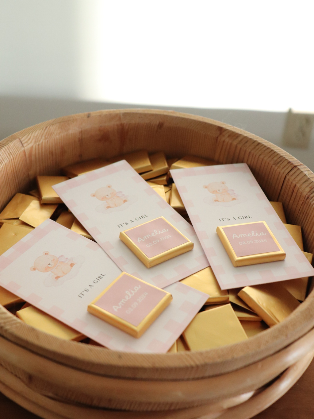 Bowl filled with small square items and cards on a light background 3 pink baby shower chocolate favours in a wooden bowl