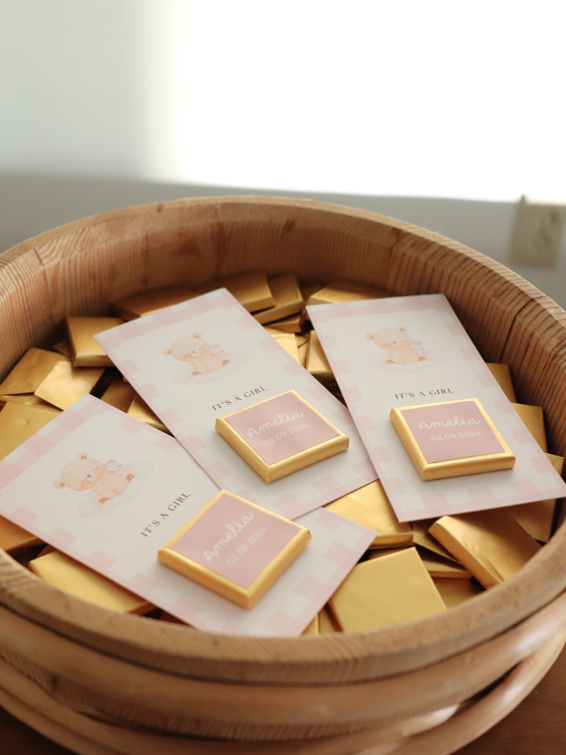 Bowl filled with small square items and cards on a light background 3 pink baby shower chocolate favours in a wooden bowl