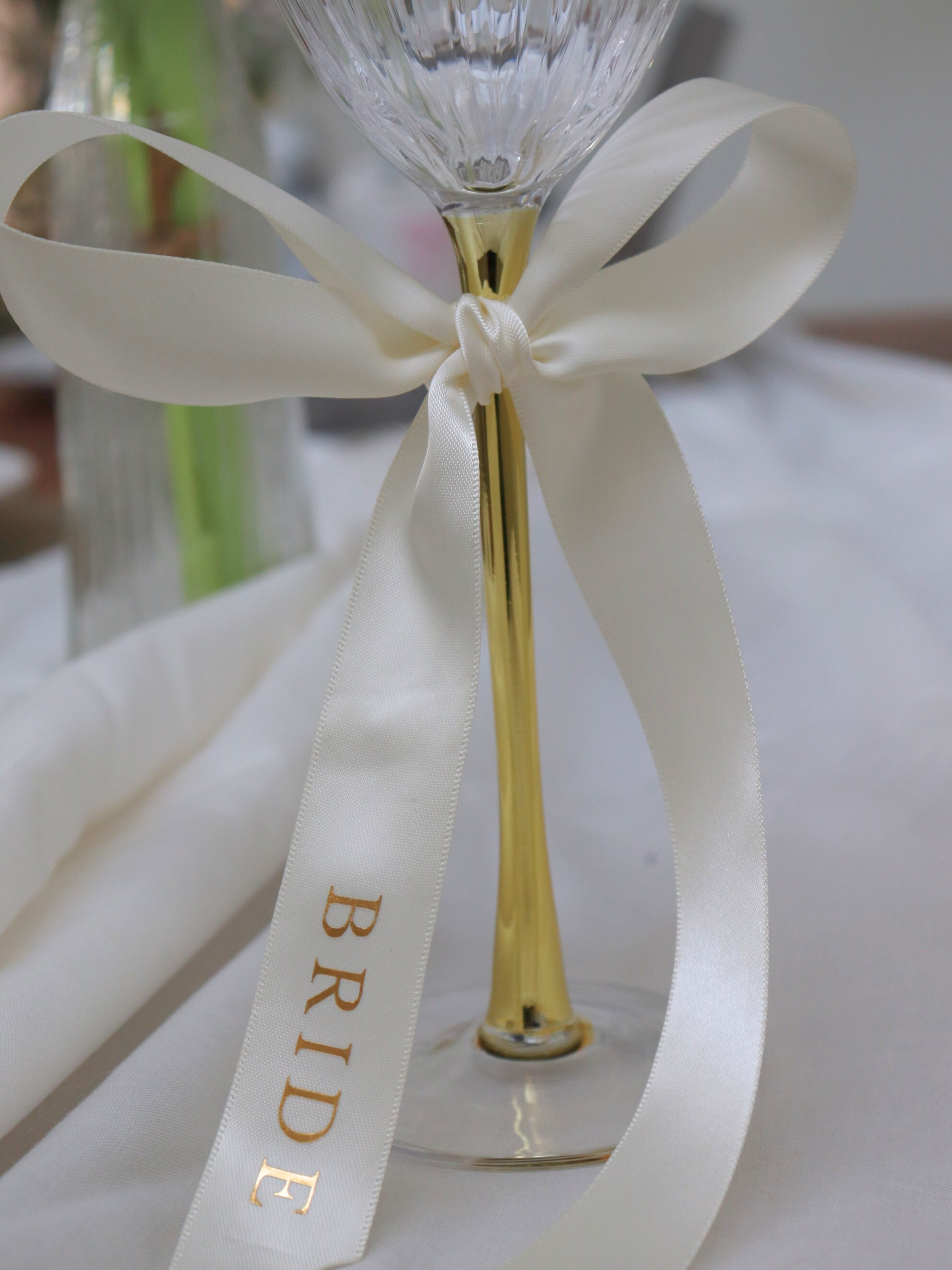 Wine glass with gold stem and white ribbon labeled 'BRIDE' on a blurred background bride gold text white personalized ribbon for wedding