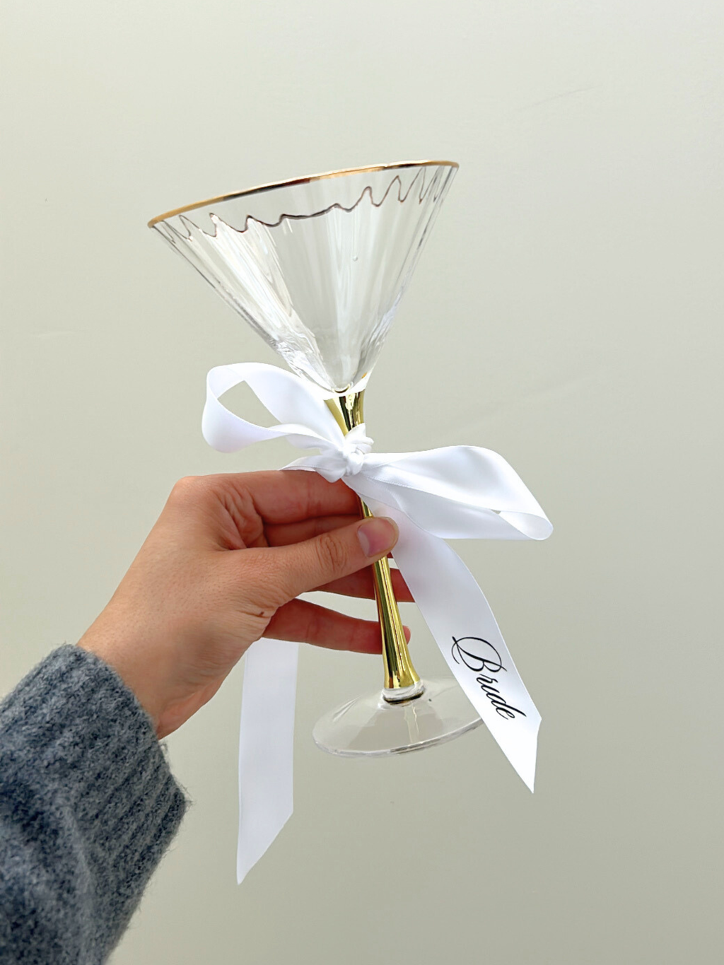 Clear martini glass with gold rim and stem, tied with a white ribbon, held by a hand against a light background. bride personalized custom wedding ribbons black and white
