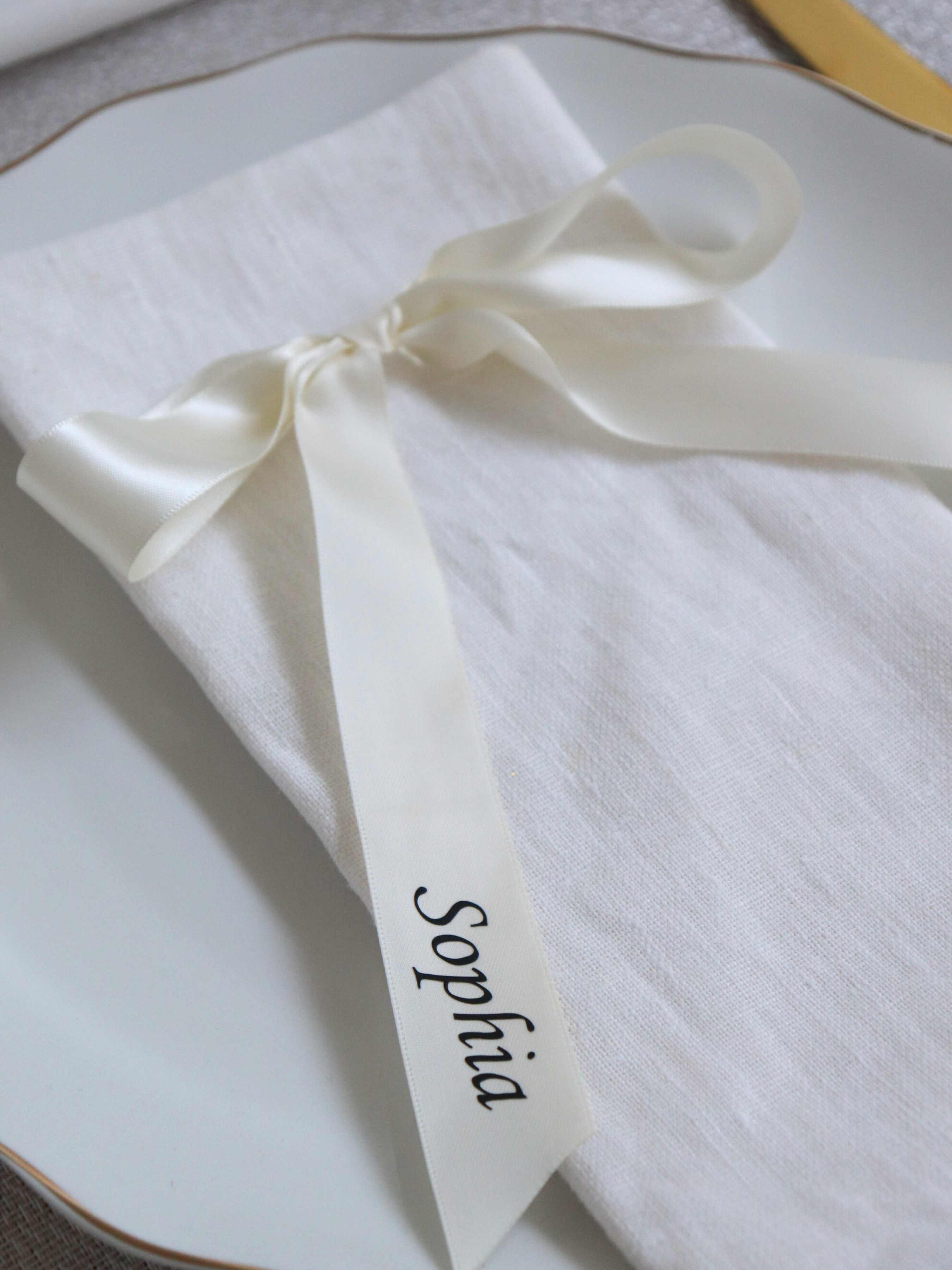 White napkin with a ribbon and 'Sophia' embroidered on it, placed on a textured surface. close up ivory personalized wedding ribbon place names for wedding