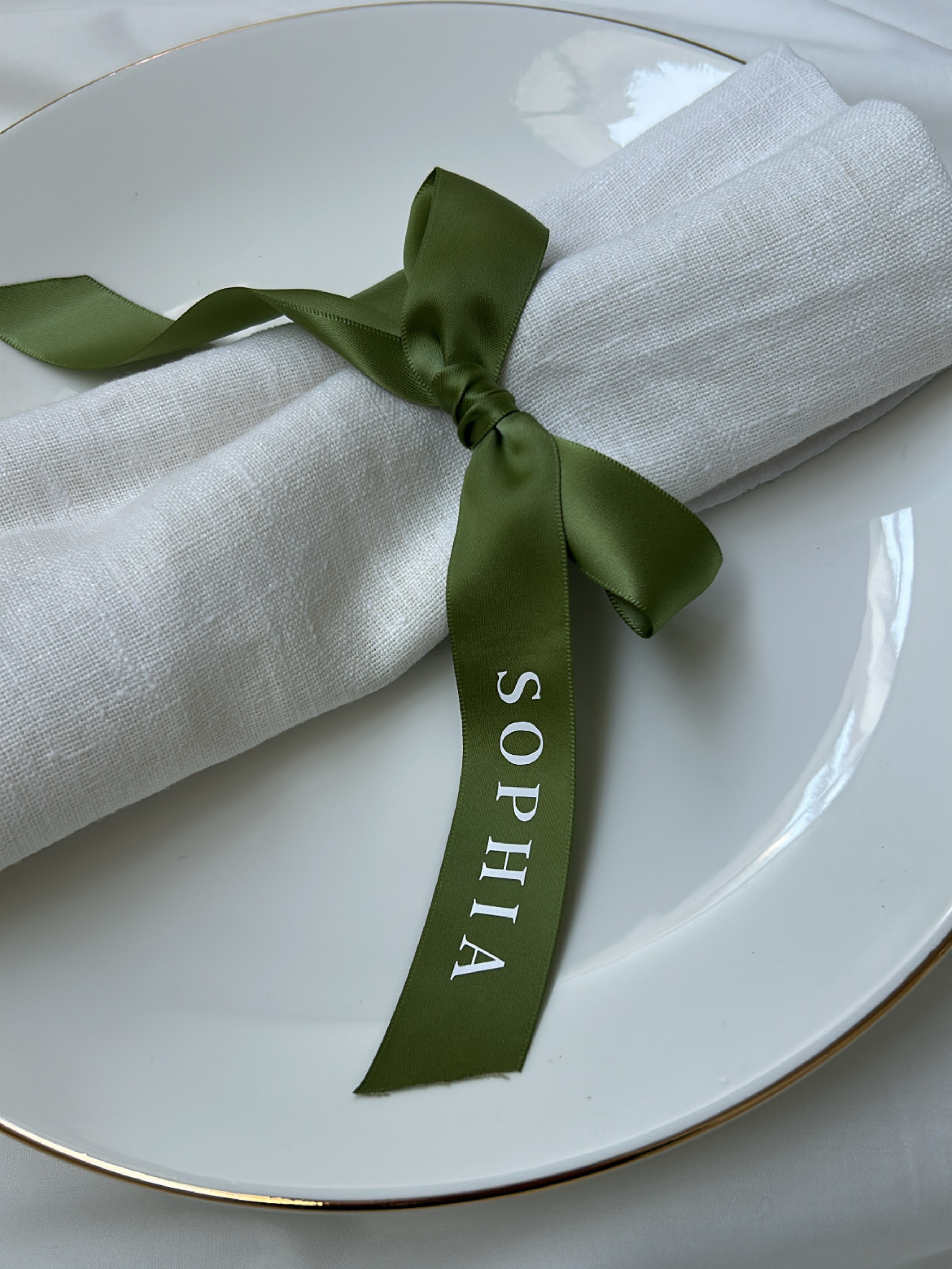 White napkin with a green ribbon tied around it on a white plate. close up ivory personalized wedding ribbon place names for wedding