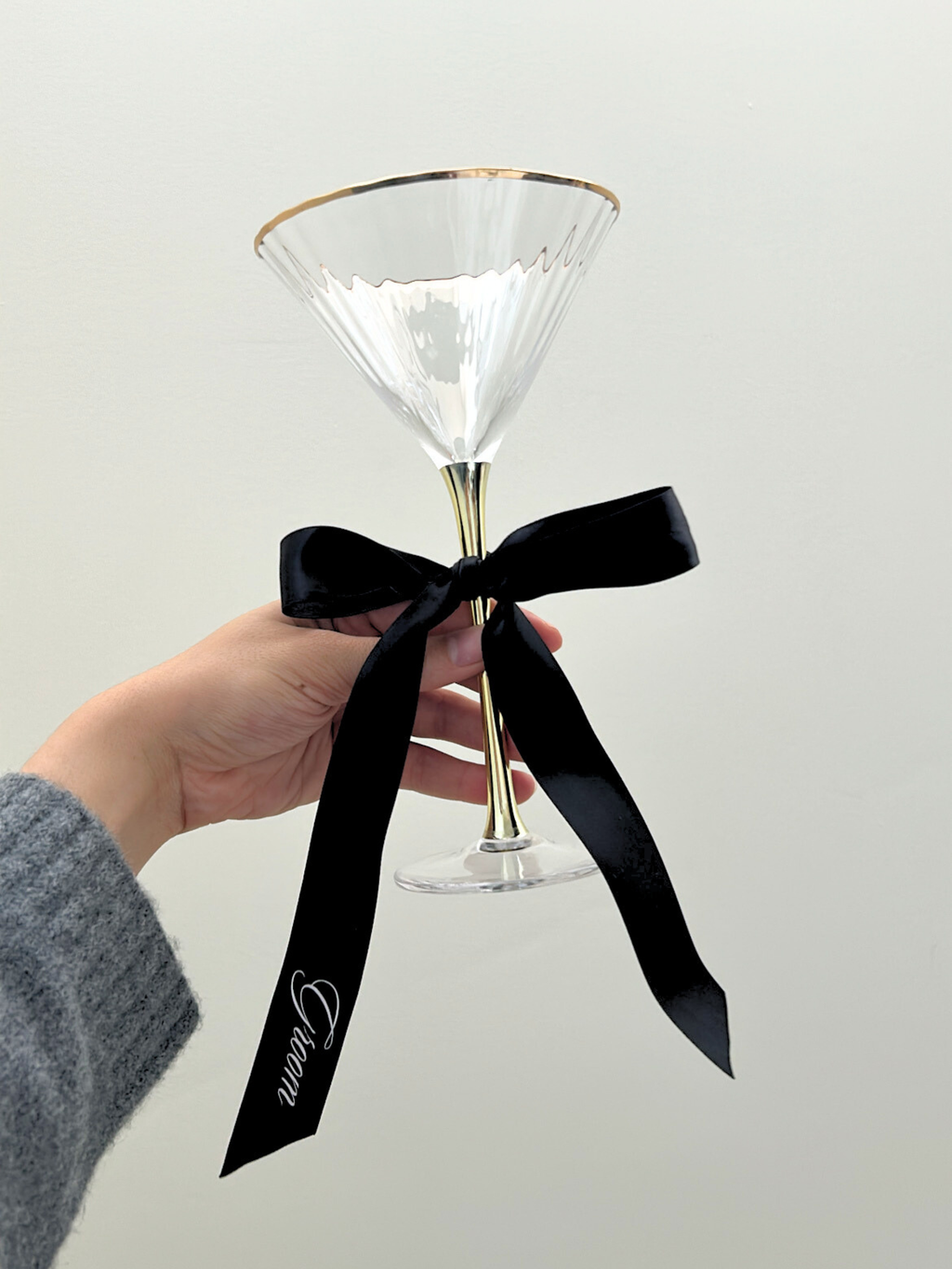Clear cocktail glass with gold rim and black ribbon held by a hand against a white background. groom personalized custom wedding ribbons black and white