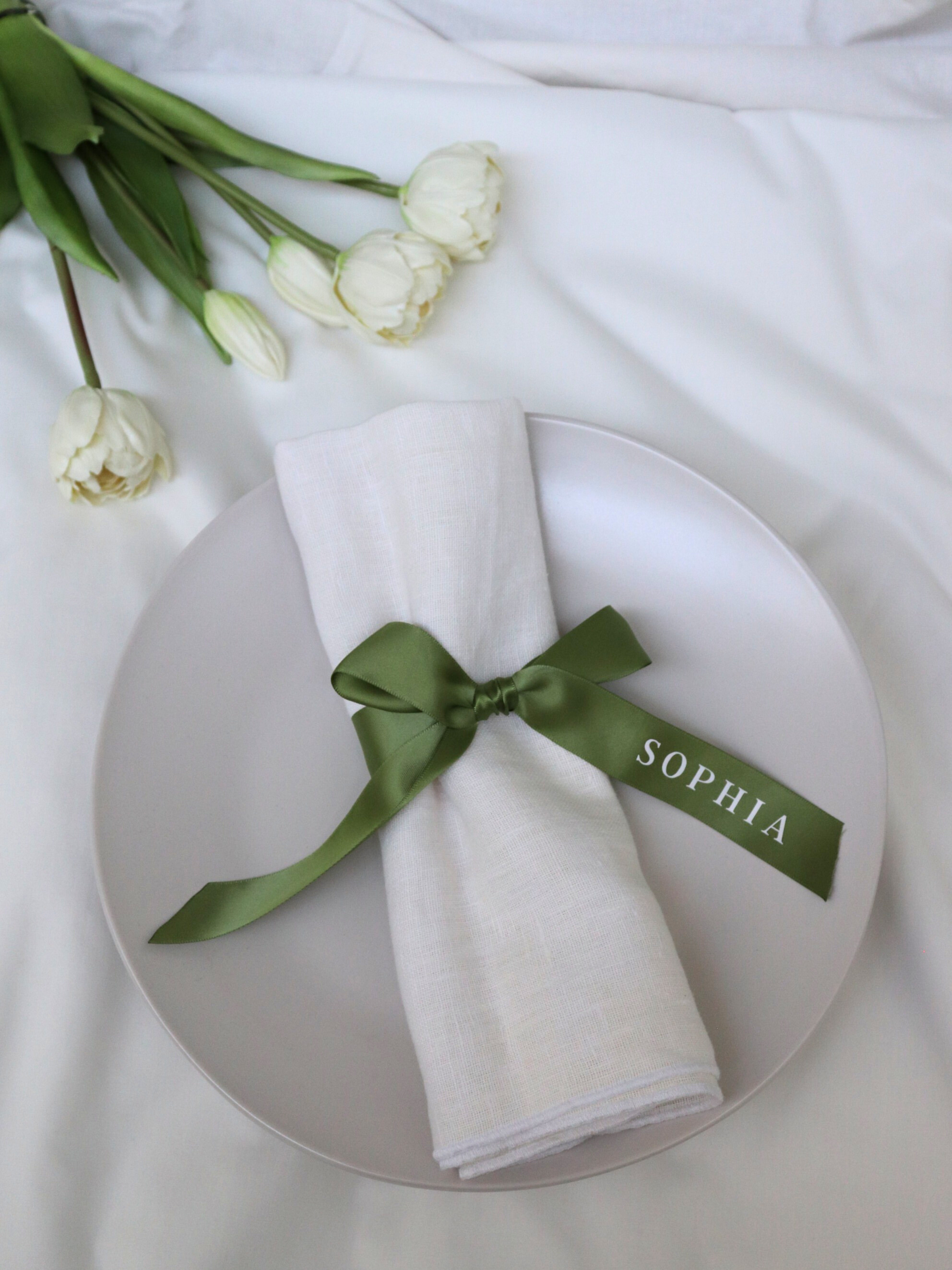 White plate with a white napkin tied with a green ribbon labeled 'SOPHIA', accompanied by white tulips on a white background.
