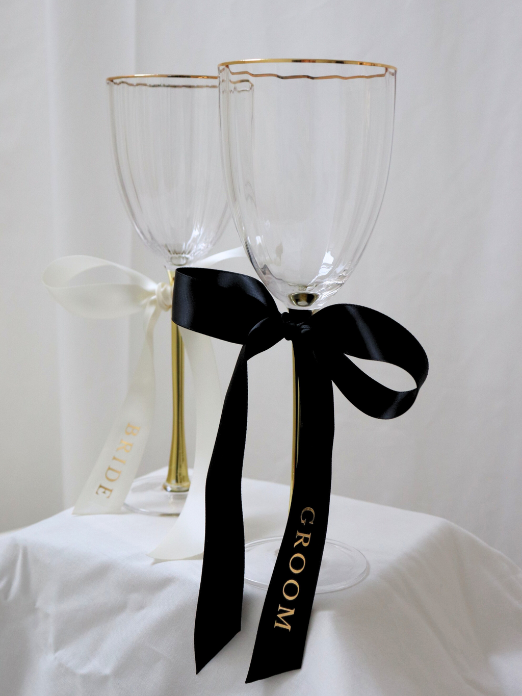Two clear wine glasses with gold rims tied together with a black ribbon on a white background. wedding gold text bride and groom personalized custom wedding ribbons black and white on white stand