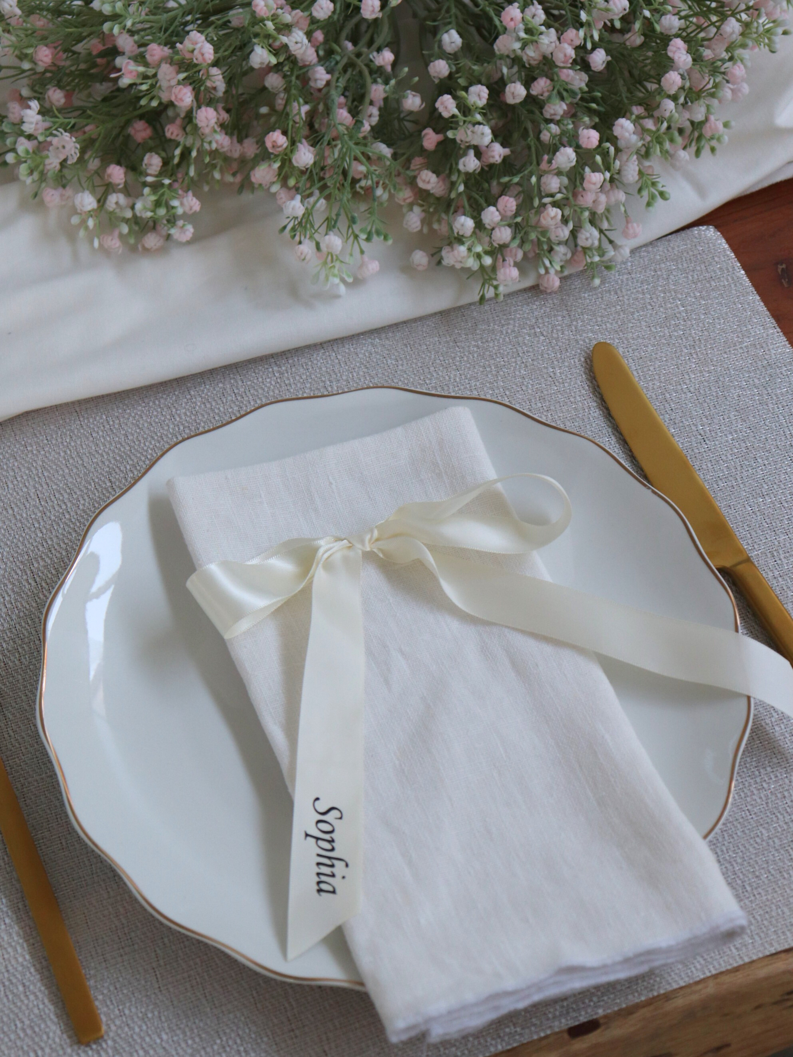 White plate with a folded napkin tied with a ribbon labeled 'Sophia', set on a table with flowers in the background. white ivory ribbon Personalized Ribbon Place Names for wedding 