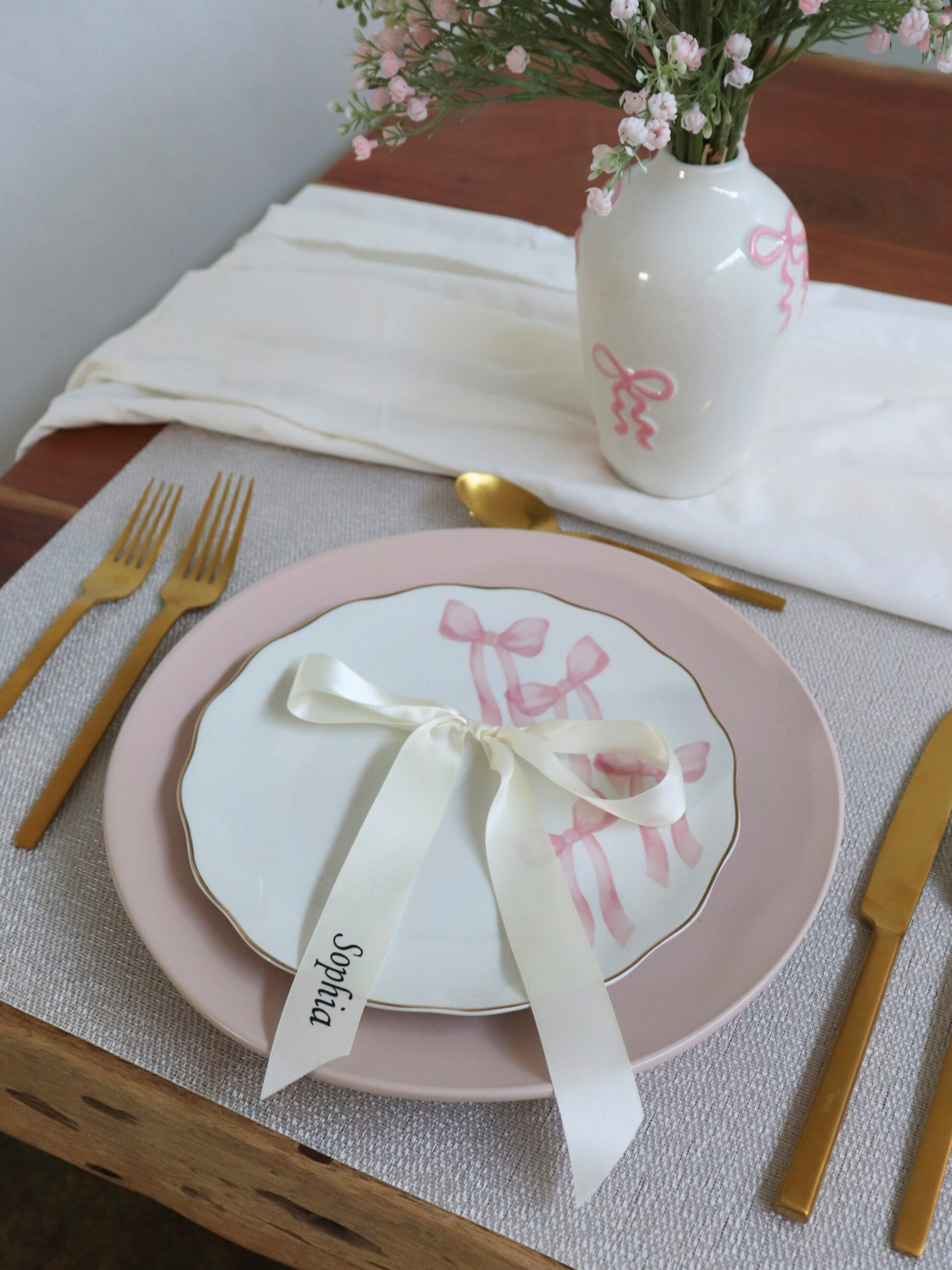 Table setting with pink plates, white napkin, and decorative elements on a wooden table. white ivory ribbon Personalized Ribbon Place Names for wedding on pink plate